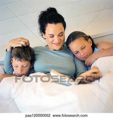 Stock Image - Mother lying in bed between children, reading book. Fotosearch