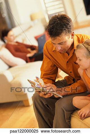 Father and daughter looking at book, woman sitting on sofa in background View Large Photo Image Stock Photography - Father and daughter looking at book, woman sitting on sofa in background. Fotosearch