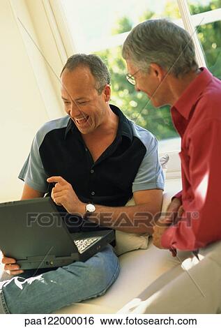 Two men sitting, one using laptop computer View Large Photo Image Stock Photograph - Two men sitting, one using laptop computer. Fotosearch