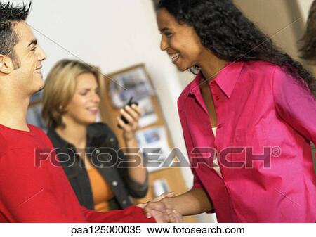 Man and woman smiling and shaking hands, woman holding cell phone in background View Large Photo Image Stock Photography - Man and woman smiling and shaking hands, woman holding cell phone in background. Fotosearch