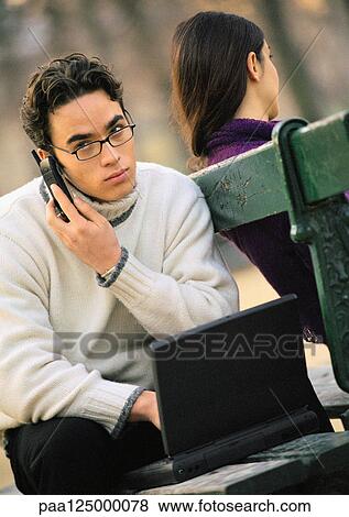 Man sitting on park bench with cell phone and laptop computer, woman sitting on other side of bench View Large Photo Image Stock Photo - Man sitting on park bench with cell phone and laptop computer, woman sitting on other side of bench. Fotosearch