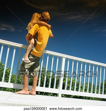 Stock Image - Boy holding little girl next to railing, rear view. Fotosearch
