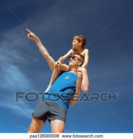 Stock Photograph - Boy sitting on man's shoulders, man pointing, outside, low angle view. Fotosearch