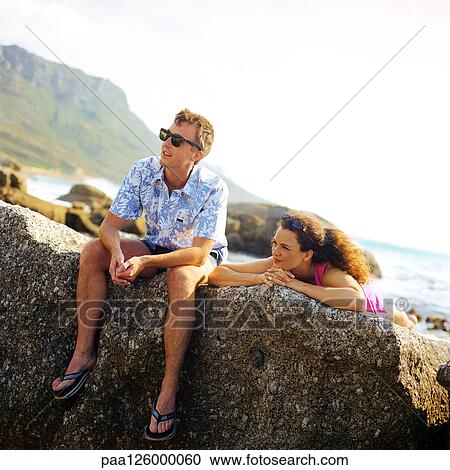 Stock Image - Couple resting on rock, sea in background. Fotosearch