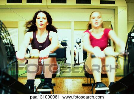 Stock Image - Women using rowing machines in gym, front view. Fotosearch