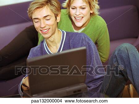 Picture - Couple smiling, man using laptop, woman sitting, looking over man's shoulder.. Fotosearch