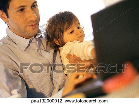 Stock Image - Father and baby, father using laptop, baby sitting on father's lap.. Fotosearch
