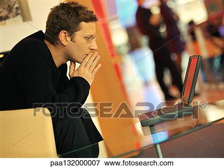 Stock Photo - Man using laptop, hands in front of mouth, side view.. Fotosearch