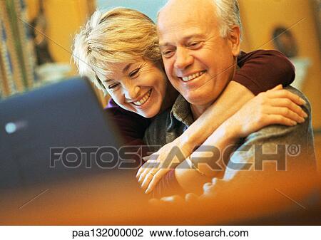 Stock Image - Mature couple smiling, man using laptop, woman with arms around man.. Fotosearch