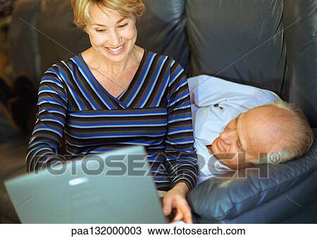 Stock Image - Mature couple, woman using laptop, man lying down.. Fotosearch