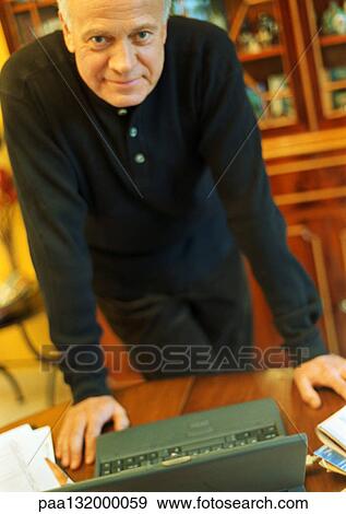 Stock Photo - Mature man leaning on desk with laptop, looking into camera.. Fotosearch