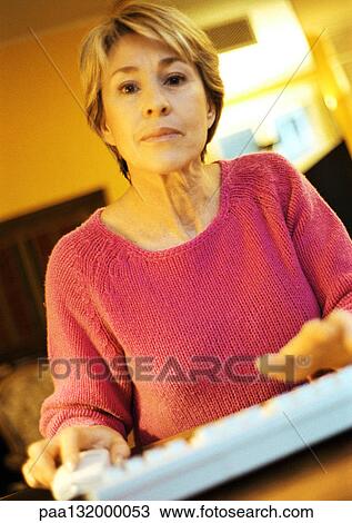 Stock Image - Mature woman, hands on computer keyboard, looking into camera, portrait.. Fotosearch