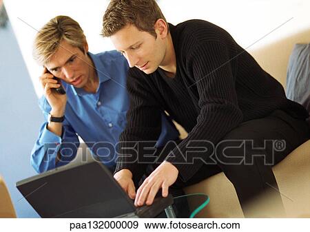 Two men, one using laptop, one using phone. View Large Photo Image Stock Photo - Two men, one using laptop, one using phone.. Fotosearch