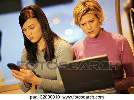 Stock Image - Two young women, one using laptop, one using phone.. Fotosearch