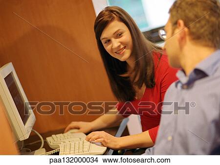 Picture - Woman using computer, man in foreground talking to her, focus on woman.. Fotosearch