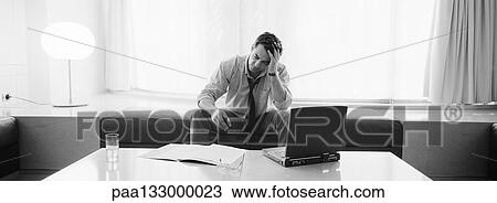 Businessman sitting holding head, laptop, drink and documents on table in front of him, b&w, panoramic view View Large Photo Image Stock Image - Businessman sitting holding head, laptop, drink and documents on table in front of him, b&w, panoramic view. Fotosearch
