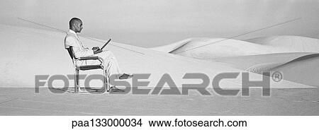 Picture - Businessman sitting in chair on dunes using laptop, b&w, panoramic view. Fotosearch