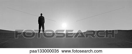 Stock Photography - Man standing on sand, backlit by sun, b&w, panoramic view. Fotosearch
