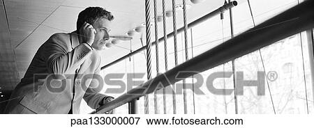 Stock Photo - Man talking on cell phone, holding onto railing and looking through window, b&w, panoramic view. Fotosearch