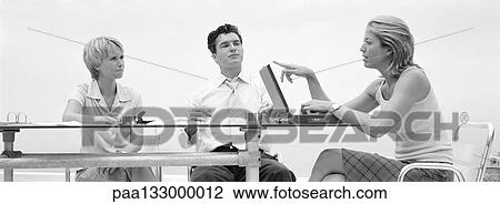 Three people sitting at table, one talking and pointing to laptop screen, b&w, panoramic view View Large Photo Image Stock Image - Three people sitting at table, one talking and pointing to laptop screen, b&w, panoramic view. Fotosearch