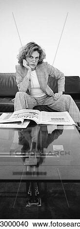 Stock Image - Woman sitting on sofa with newspaper open on coffee table, looking at camera, b&w, vertical. Fotosearch