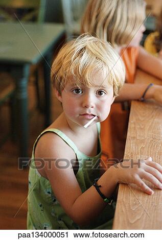 Stock Image - Little girl sitting at table with other children, sucking lollipop, looking into camera.. Fotosearch