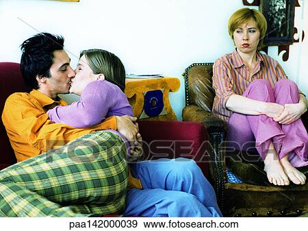 Stock Photo - Couple on sofa kissing, second woman sitting in armchair, watching.. Fotosearch
