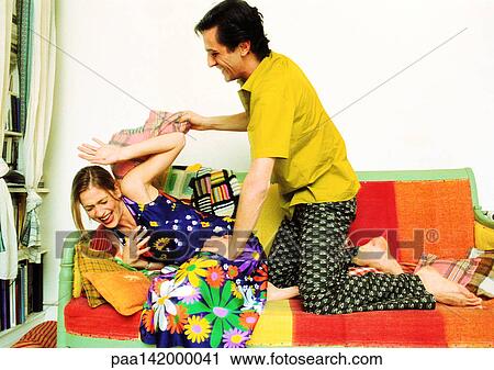 Stock Image - Couple on sofa, man hitting woman with cushion, woman laughing.. Fotosearch