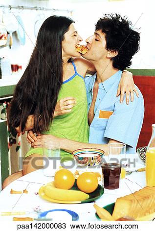 Couple sitting at breakfast table, woman sitting on man's lap, eating from ends of same croissant View Large Photo Image Stock Image - Couple sitting at breakfast table, woman sitting on man's lap, eating from ends of same croissant. Fotosearch