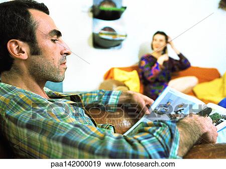 Stock Photo - Man reading magazine, woman sitting in background.. Fotosearch