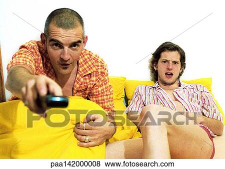 Stock Photo - Two men sitting on sofa, one pointing remote control at camera. Fotosearch