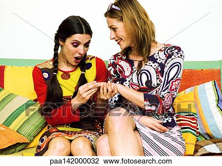 Stock Image - Two young women sitting on sofa, one showing other hand.. Fotosearch