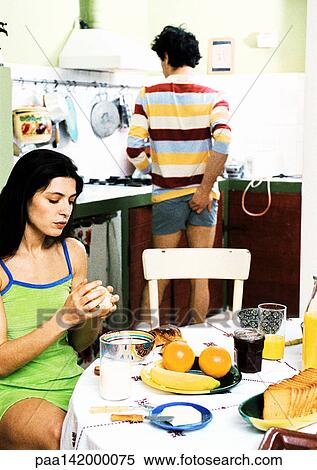 Stock Photography - Woman sitting at breakfast table, man standing, scratching rear. Fotosearch