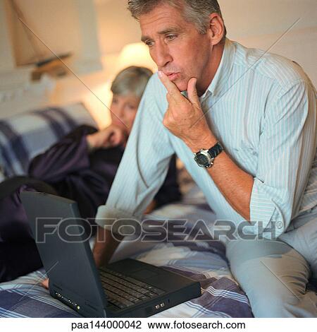 Mature man using laptop computer on bed, woman in background View Large Photo Image Stock Image - Mature man using laptop computer on bed, woman in background. Fotosearch