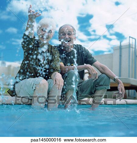 Stock Photograph - Mature couple sitting on edge of swimming pool, woman splashing water into air. Fotosearch