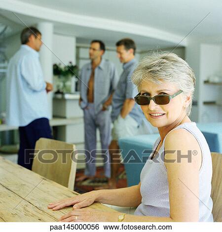 Stock Photograph - Mature woman sitting at table, three men standing in background. Fotosearch