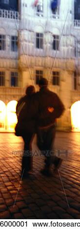 Stock Image - Couple walking with arms around each other at night, rear view, blurred. Fotosearch