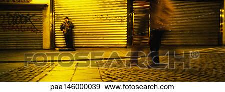 Musician leaning against roller shutter, people walking on cobble stone street, blurred motion View Large Photo Image Stock Photo - Musician leaning against roller shutter, people walking on cobble stone street, blurred motion. Fotosearch