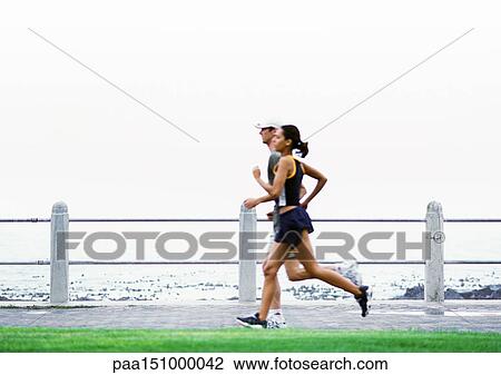 Stock Image - Couple jogging, sea in background, side view.. Fotosearch