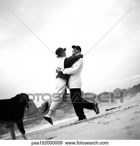 Stock Photo - Mature couple and dog on beach, woman jumping into man's arms, b&w.. Fotosearch