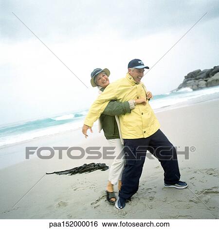 Stock Photograph - Mature couple playing on beach, woman grabbing man from behind.. Fotosearch