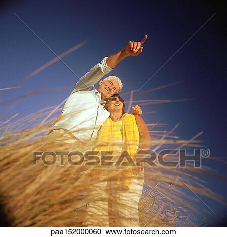 Stock Image - Mature couple standing in tall grass, low angle view.. Fotosearch
