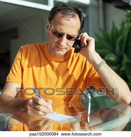 Picture - Mature man using phone, taking notes at table. Fotosearch