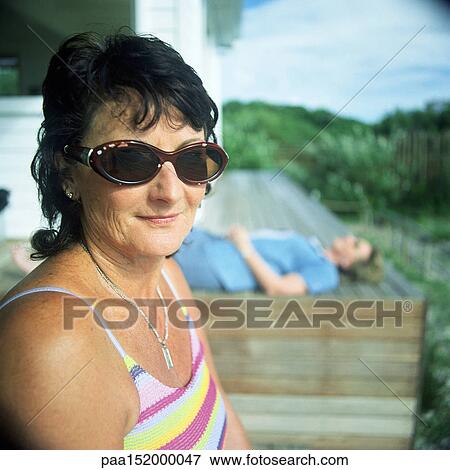 Stock Photo - Mature woman sitting on a bench, wearing sunglasses, second person lying down in background. Fotosearch