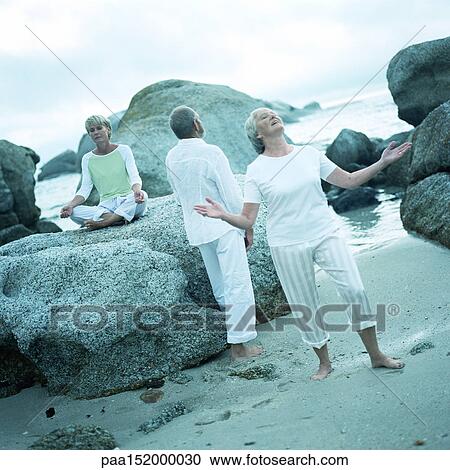 Stock Image - Three mature people in different poses on beach.. Fotosearch