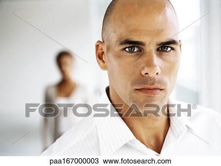 Stock Image - Man looking into camera, woman in background, portrait, close-up. Fotosearch