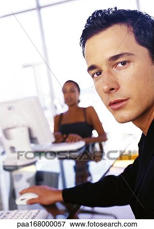 Stock Photo - Businessman and businesswoman working at desks, man looking at camera. Fotosearch
