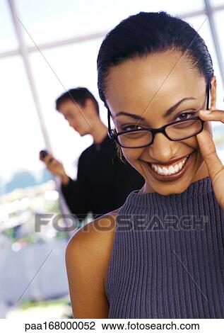 Businesswoman lowering glasses and smiling, man using phone in background, portrait View Large Photo Image Stock Image - Businesswoman lowering glasses and smiling, man using phone in background, portrait. Fotosearch