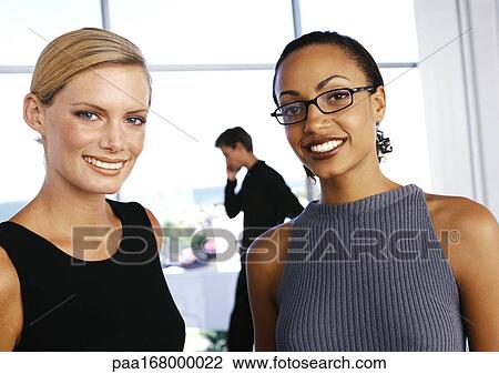 Stock Image - Businesswomen smiling, man talking on phone in background. Fotosearch