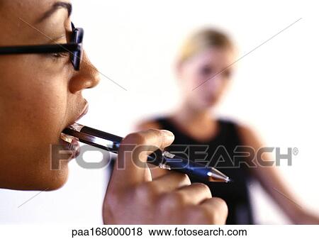 Woman holding pen in mouth, second woman blurred in background View Large Photo Image Stock Photo - Woman holding pen in mouth, second woman blurred in background. Fotosearch